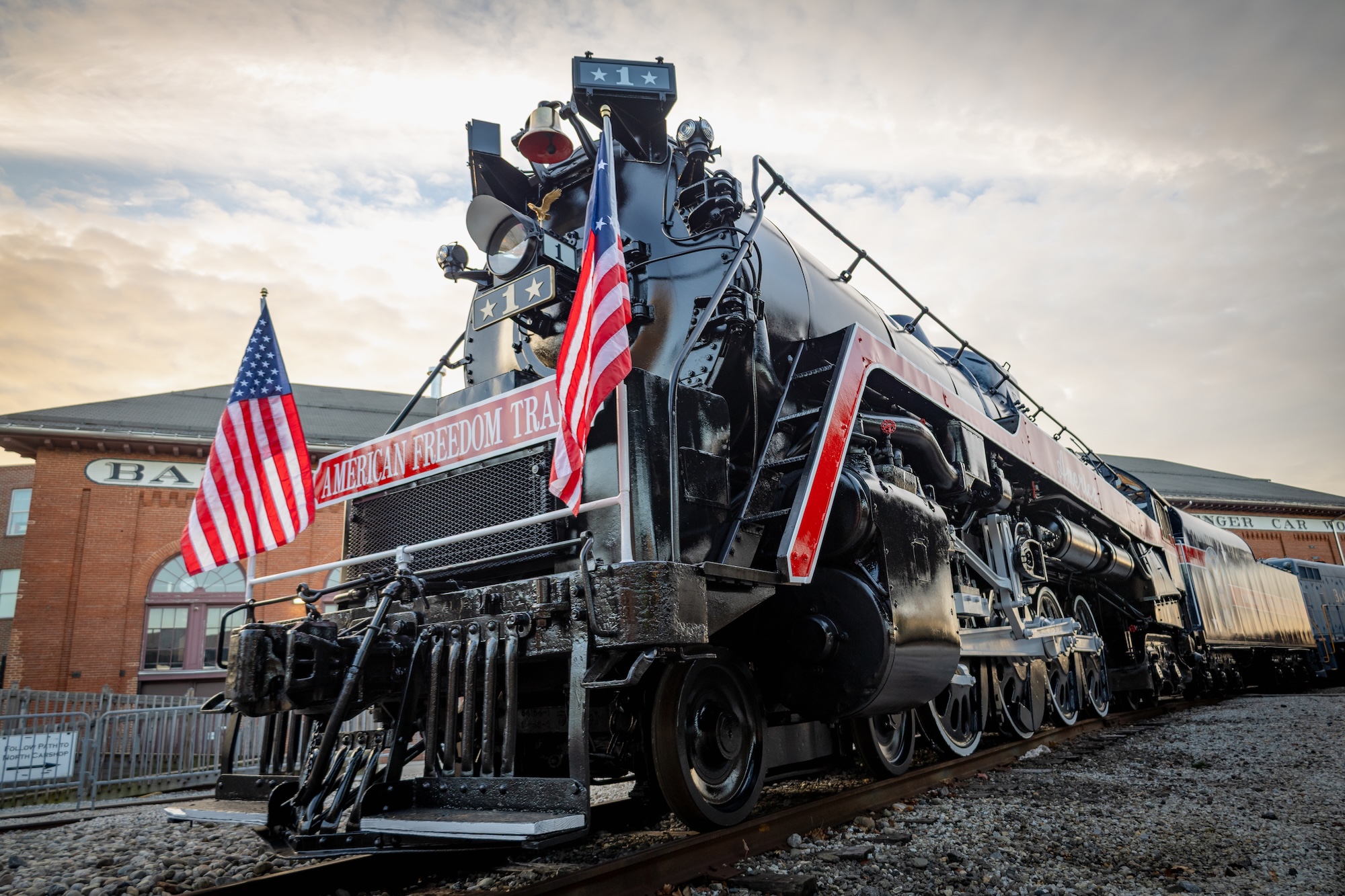 The American Freedom Train No. 1 - a large steam locomotive with two American flags flying in the front - sits outside the North Car shop of the B&O Railroad Museum.